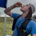 Load image into Gallery viewer, A man in a blue shirt and black hydration vest drinking from a small bottle outdoors under a blue canopy, with a cloudy sky in the background.
