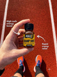 Load image into Gallery viewer, A hand holds a small bottle of PickleUp 'Cramp Shot' food jam and tea coffee mix on an outdoor running track. The image includes text overlays describing the product's benefits: 'High carb energy boost' and 'Rapid cramp relief'.
